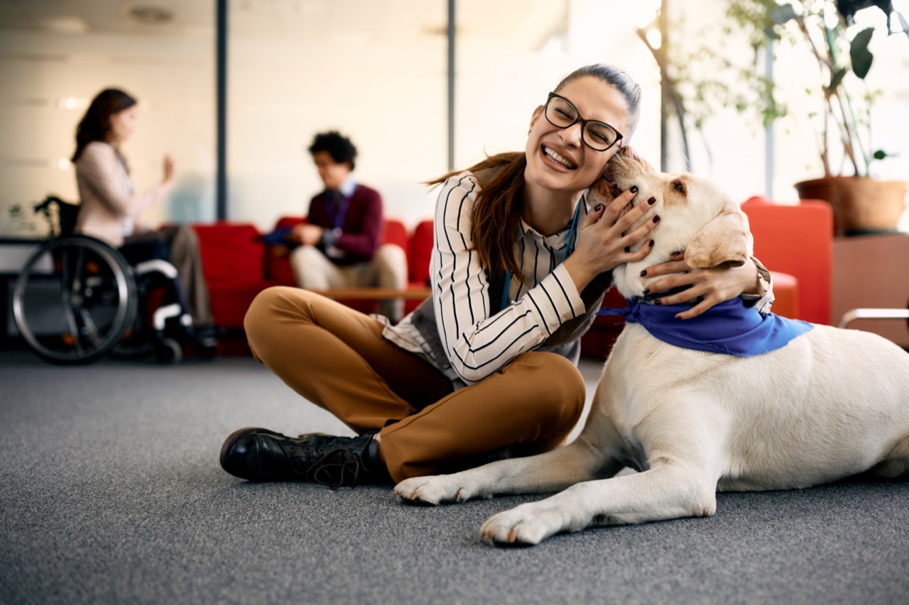 A woman laughing as her yellow Labrador service dog leans in to kiss her cheek